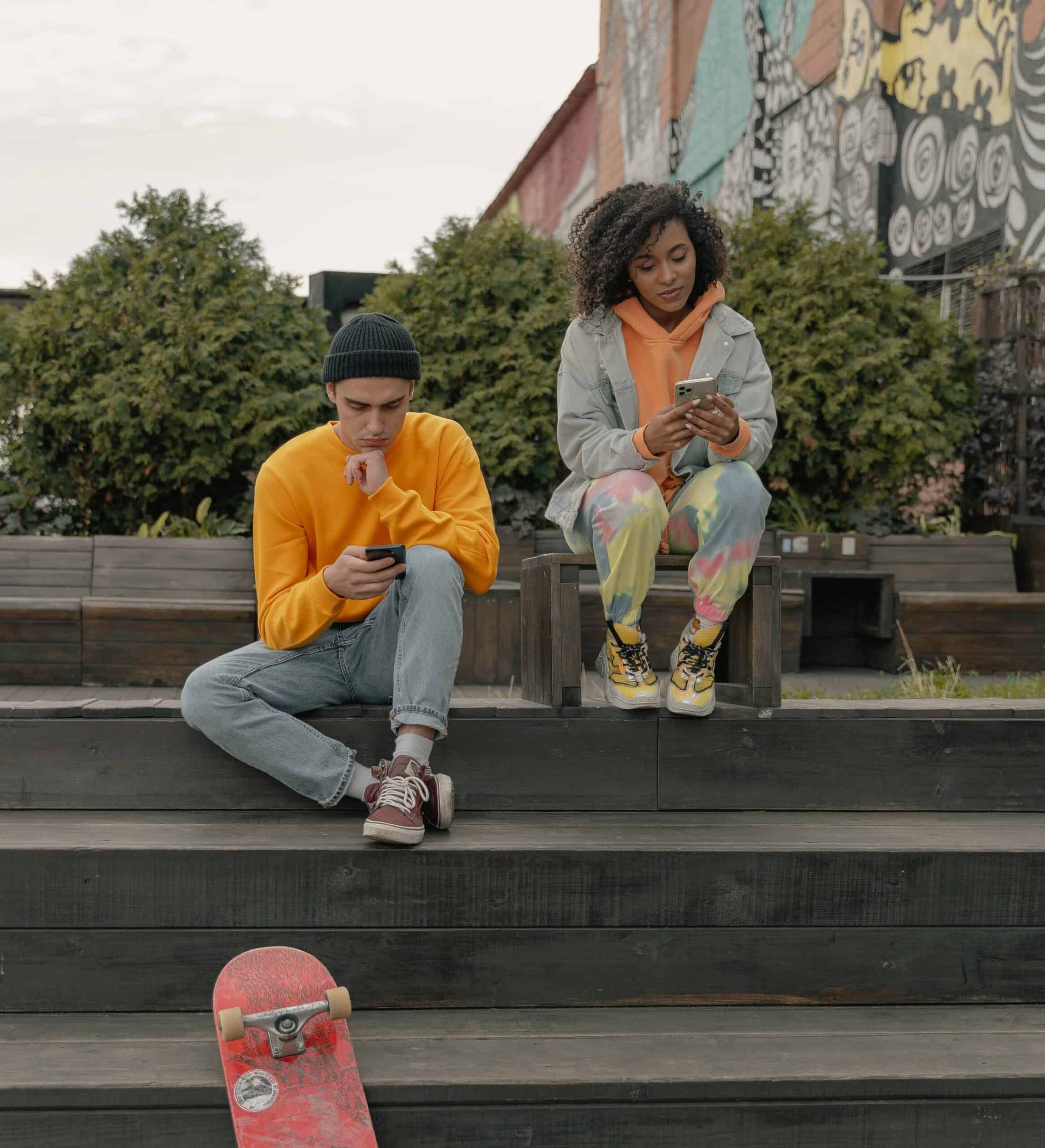Two people looking at their phones sitting on steps with a skateboard